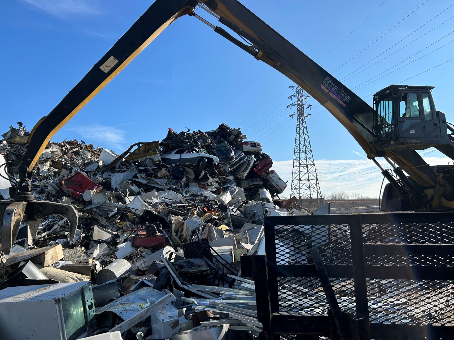 A front loader sorts scrap metal at a recycling center with a blue sky and power lines in the background.