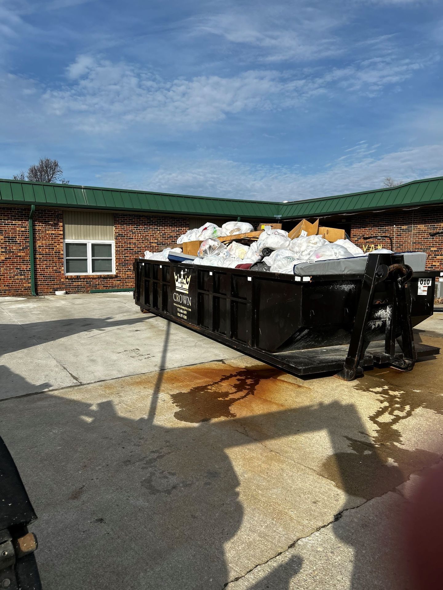 Black dumpster overflowing with trash, placed in front of a brick building under a partly cloudy blue sky.