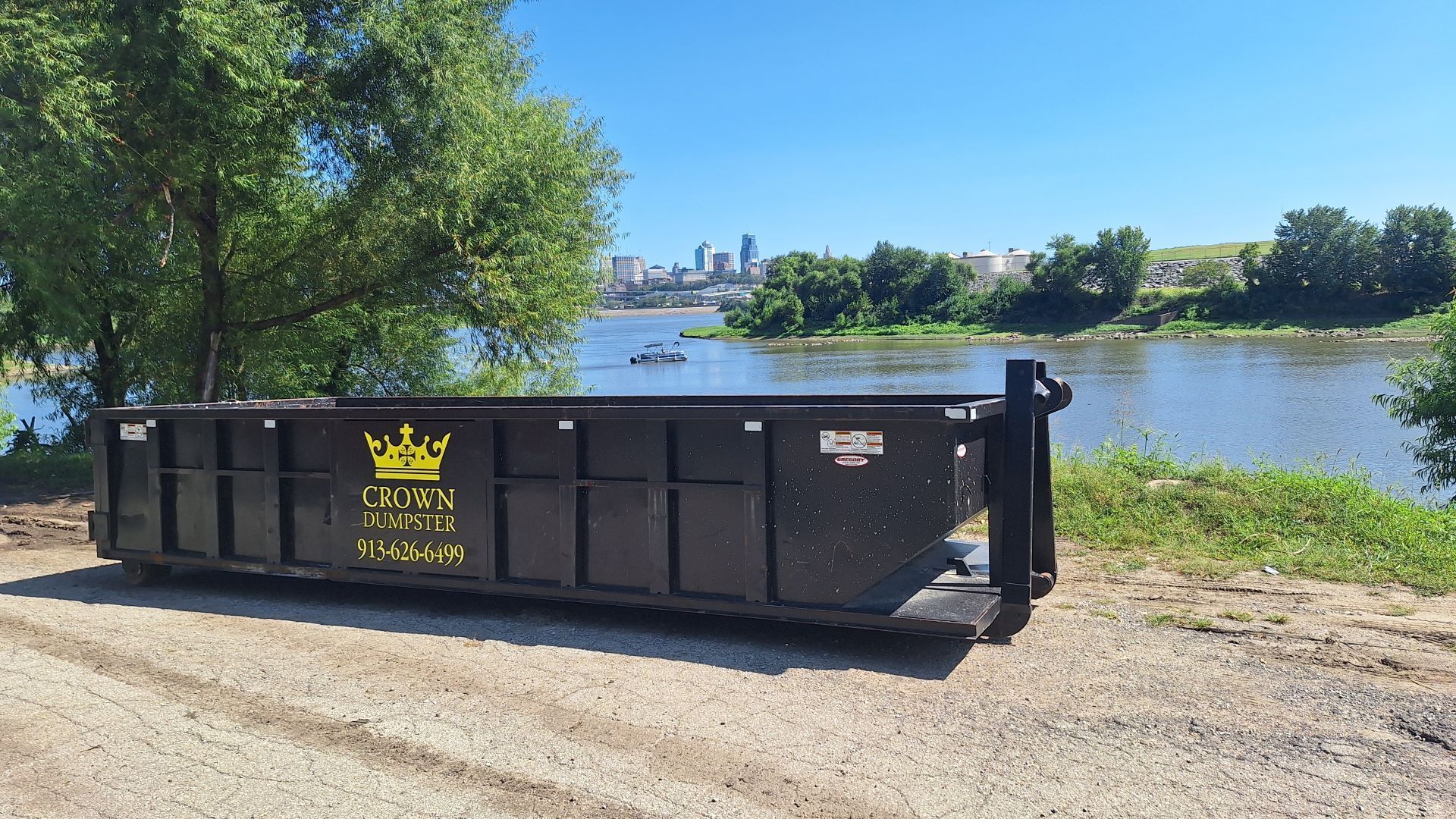 Black dumpster by a river, with city skyline in the distance on a sunny day.