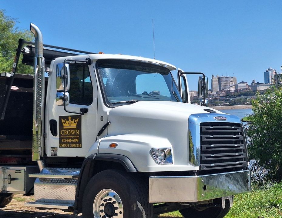 White Crown company dump truck parked near water, city skyline in the background.