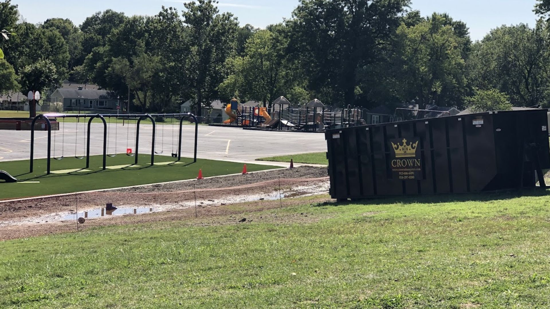 Park scene with a playground, a dumpster, and a water feature. Green grass and trees under a sunny sky.
