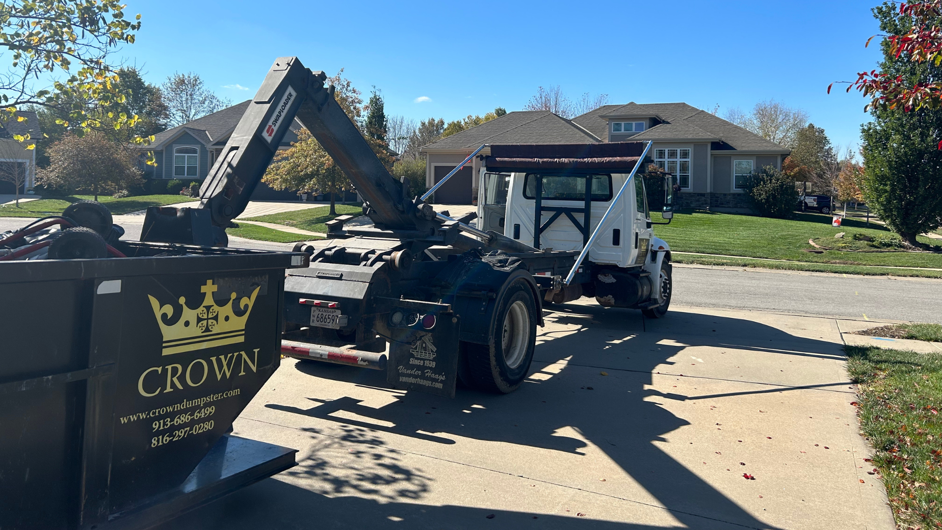 Truck with a grapple arm loading a black dumpster in a residential driveway on a sunny day.