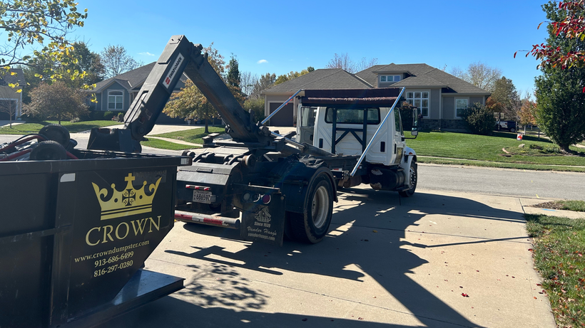 Truck with a grapple arm loading a black dumpster in a residential driveway on a sunny day.
