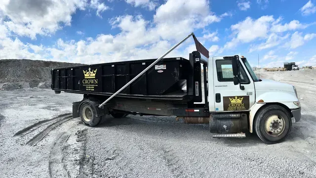 Dump truck with Crown Waste logo on a gravel surface under a cloudy sky.