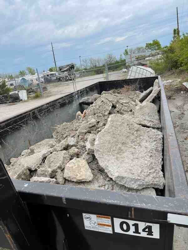Black dumpster filled with broken concrete rubble outdoors. Number 014 on side. Cloudy sky.