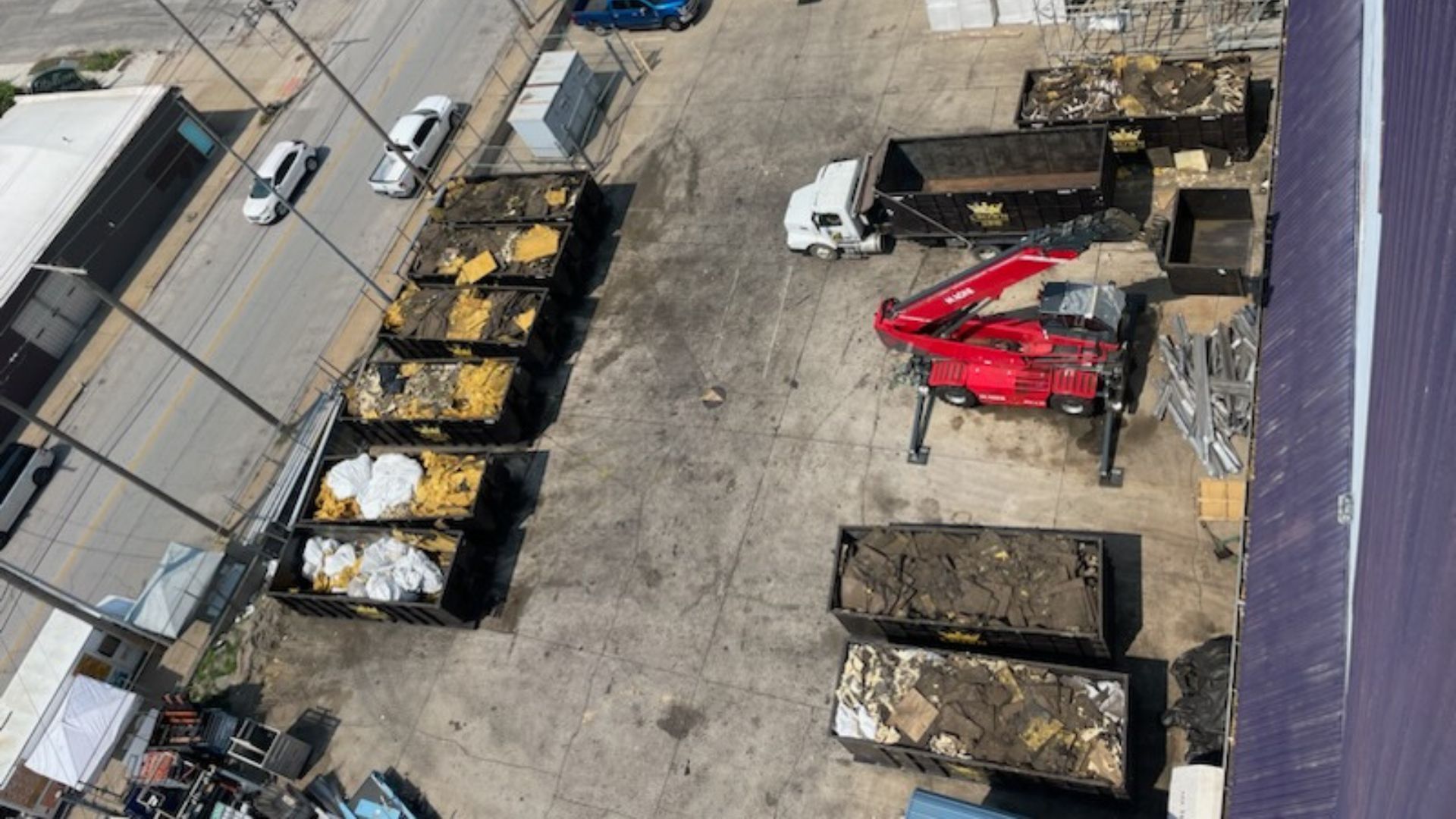 Overhead view of a debris processing area with dumpsters, a red excavator, and a truck parked on concrete.