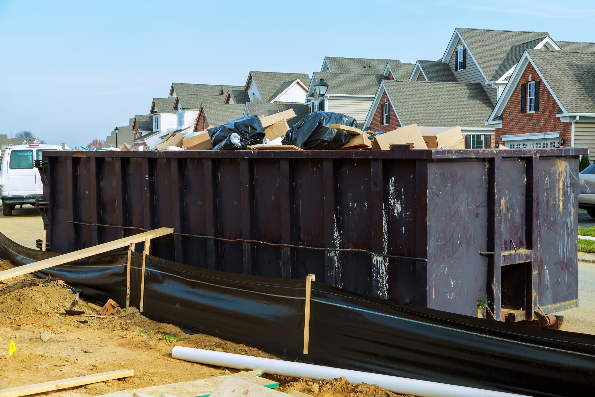 A large, dark brown construction dumpster filled with debris sits next to a residential construction site.