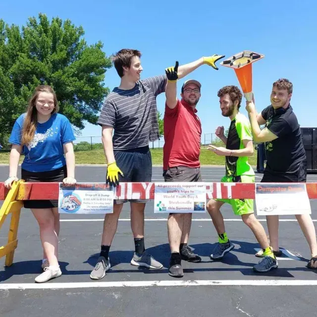 Five people posing behind a road barrier in a parking lot. Some are holding a traffic cone. Sunny day.