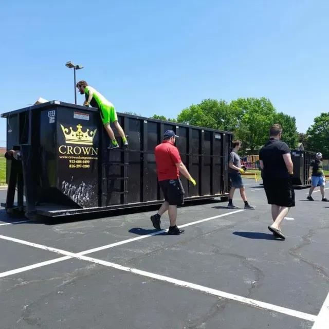 People near large dumpster, one person on top. Sunny outdoor setting.