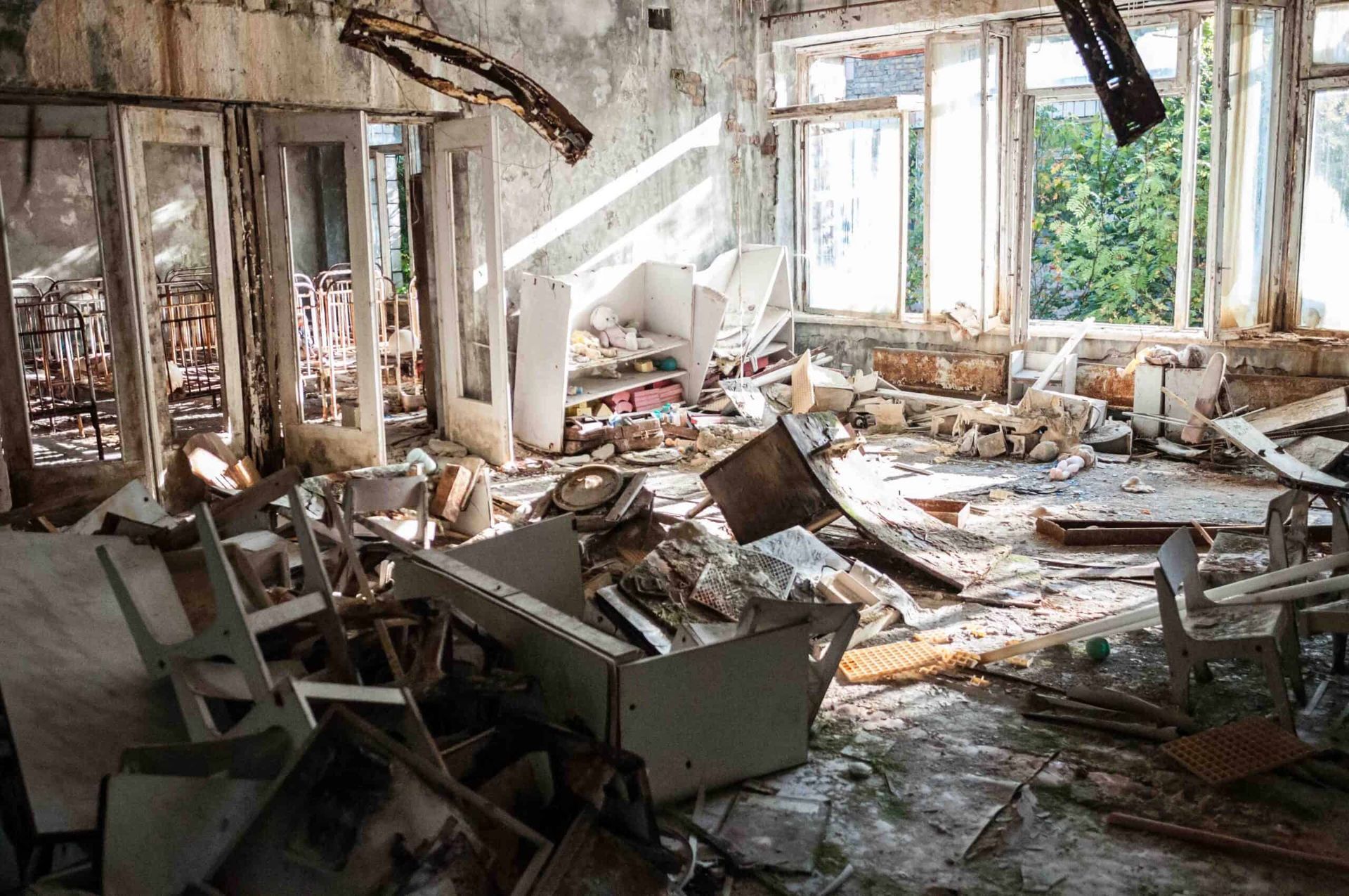 Debris-filled room with broken furniture and windows, damaged walls, in a dilapidated building.