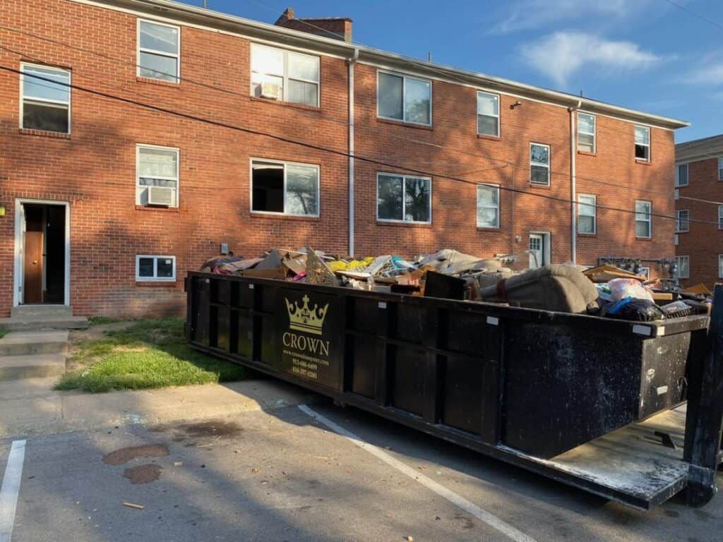 A black dumpster overflowing with debris sits in front of a brick apartment building.