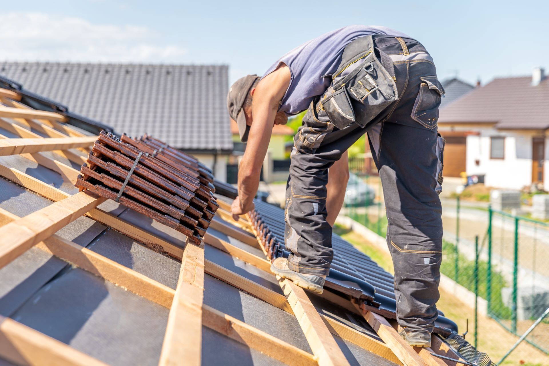 Roofer installing tiles on a residential roof, wearing work pants and a cap, on a sunny day.