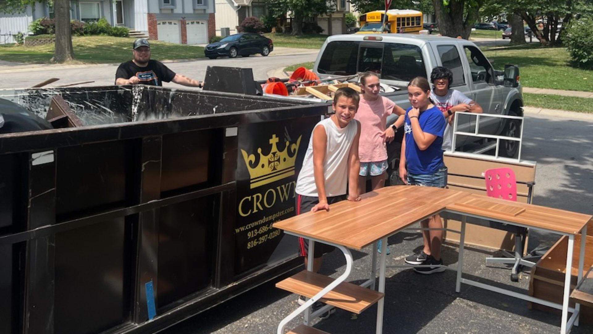 People near a dumpster, a truck, and a desk outside on a sunny day.