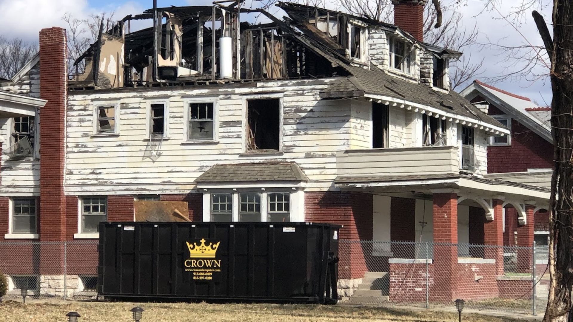Burned-out two-story house with damaged roof and a dumpster in front.