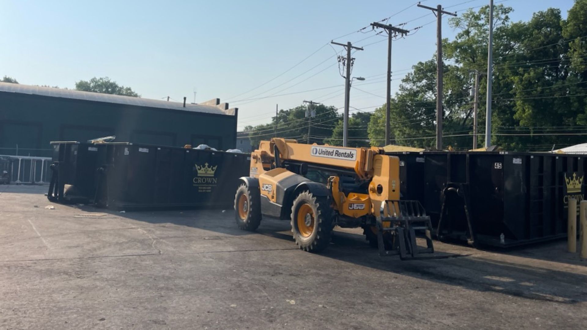 Yellow forklift moving a black dumpster in an outdoor lot on a sunny day.