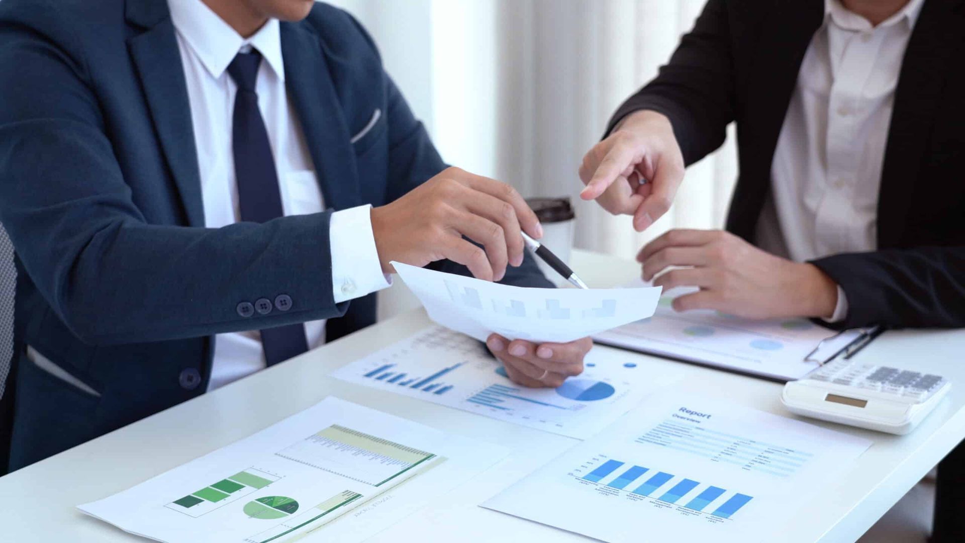 Two people in suits reviewing financial documents and pointing. Graphs and a calculator are on the desk.