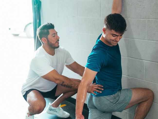 a man is helping another man stretch his legs in a gym .