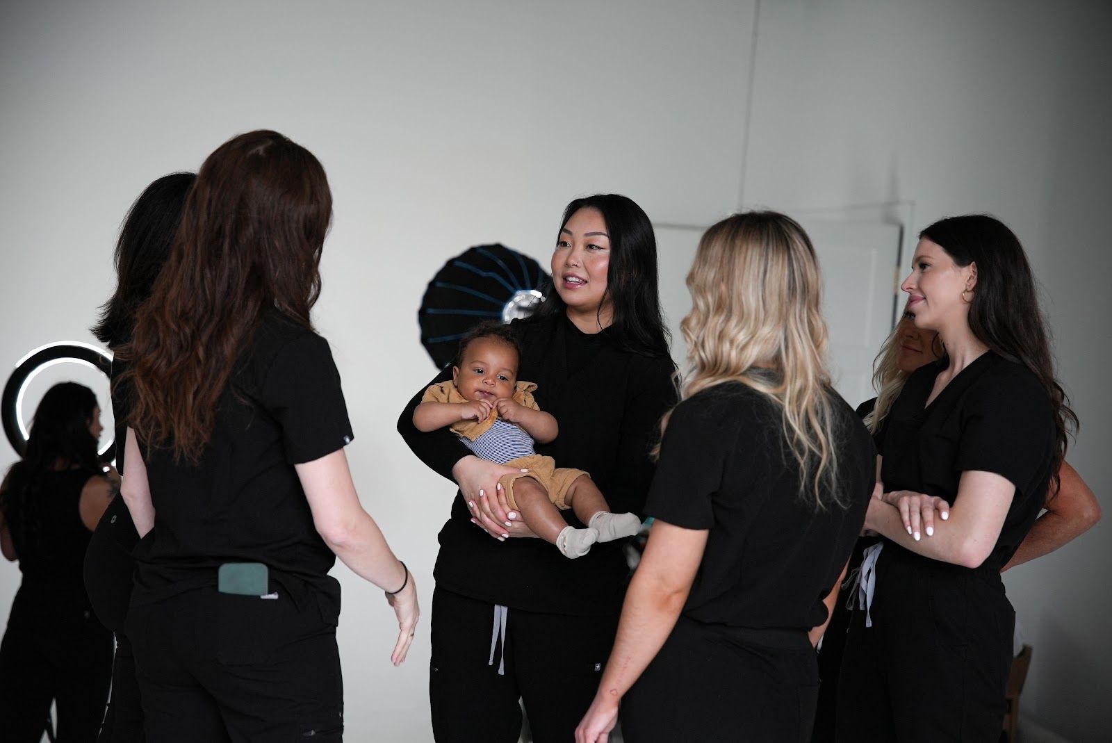 A group of people wearing black scrubs stand in a bright room, surrounding someone holding a baby.