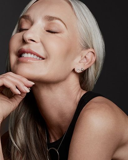 Woman with silver hair, eyes closed, smiling, touching her chin, wearing a necklace and earrings.