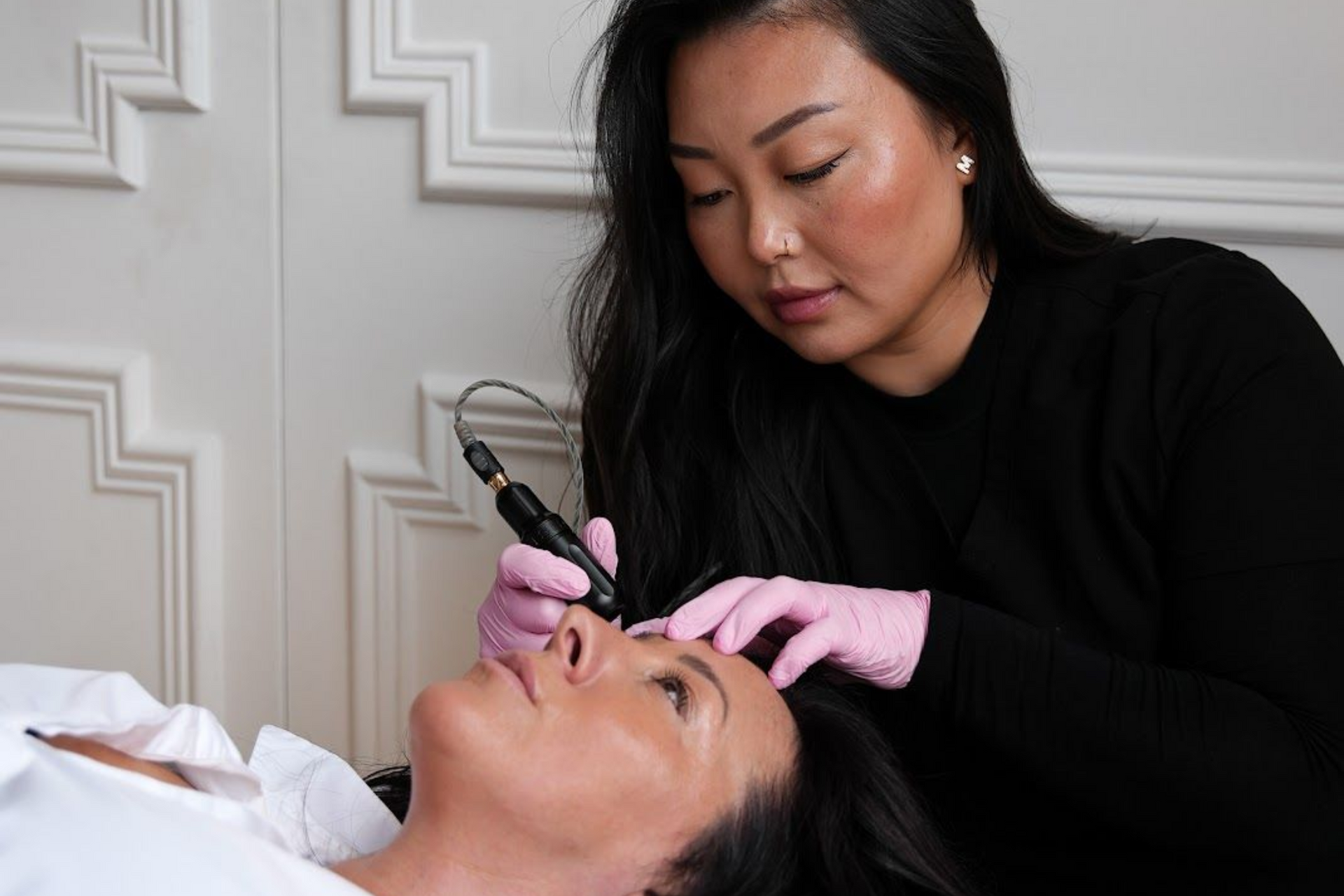 A technician wearing pink gloves uses a handheld cosmetic tattooing device on a client’s eyebrow in a studio.