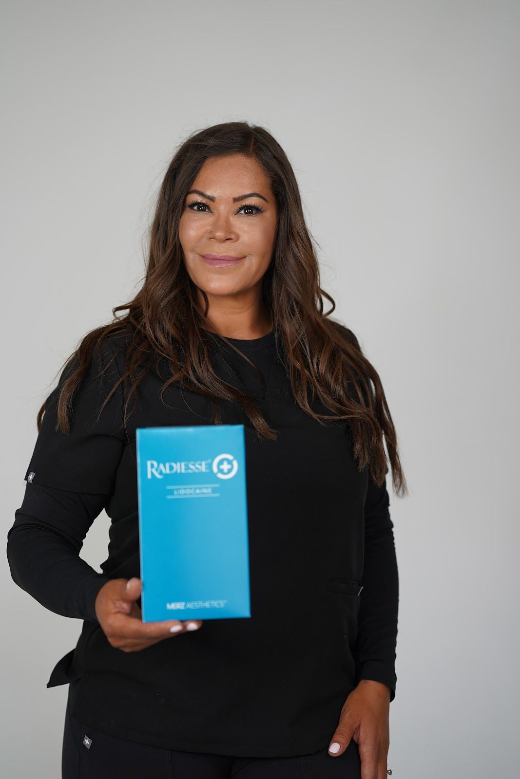 Woman in black scrubs holding a blue box with white text. Neutral background, smiling.