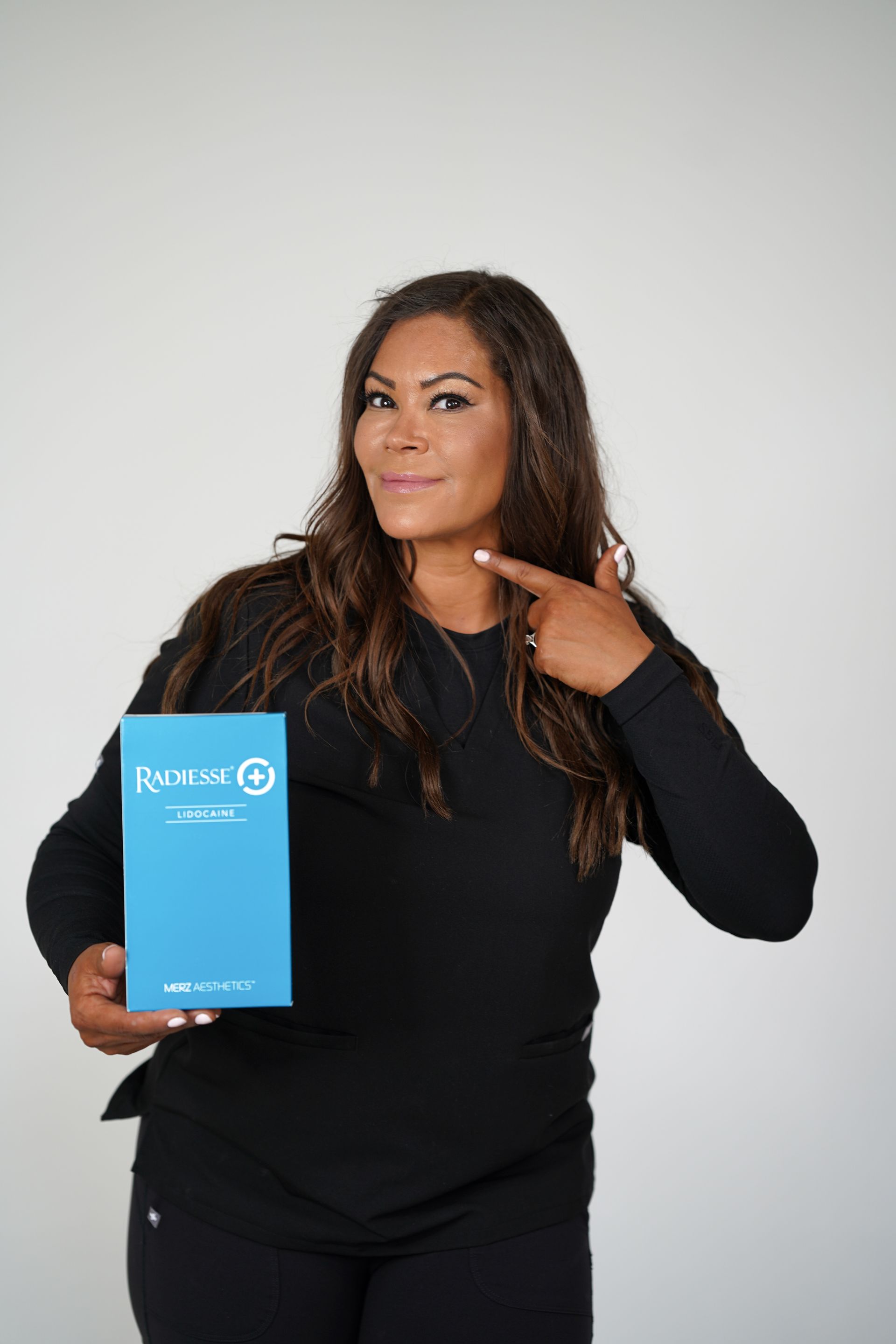 Woman in black points to her neck, holding a blue product box against a white backdrop.