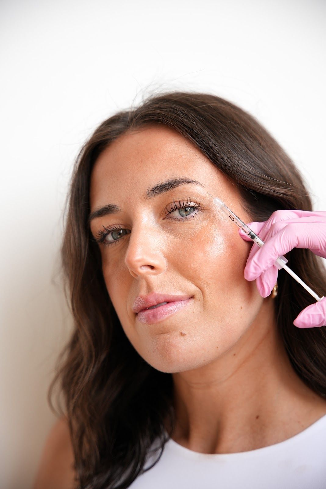 Woman receiving a facial injection, with pink-gloved hand holding a syringe near her eye, against a white background.