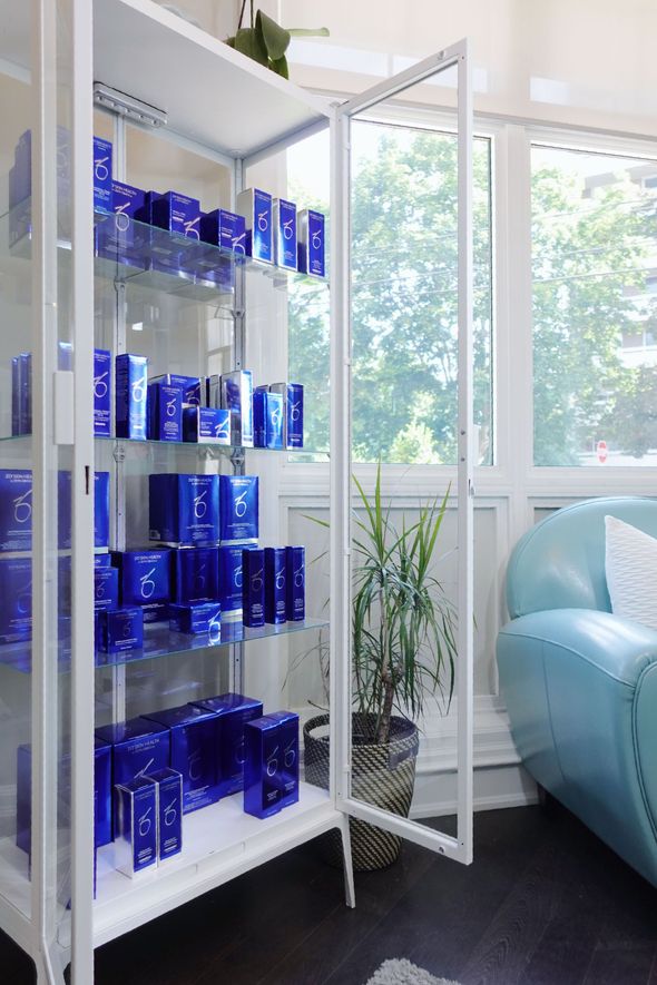 White cabinet with glass doors displaying blue skincare products, near a window and blue sofa.