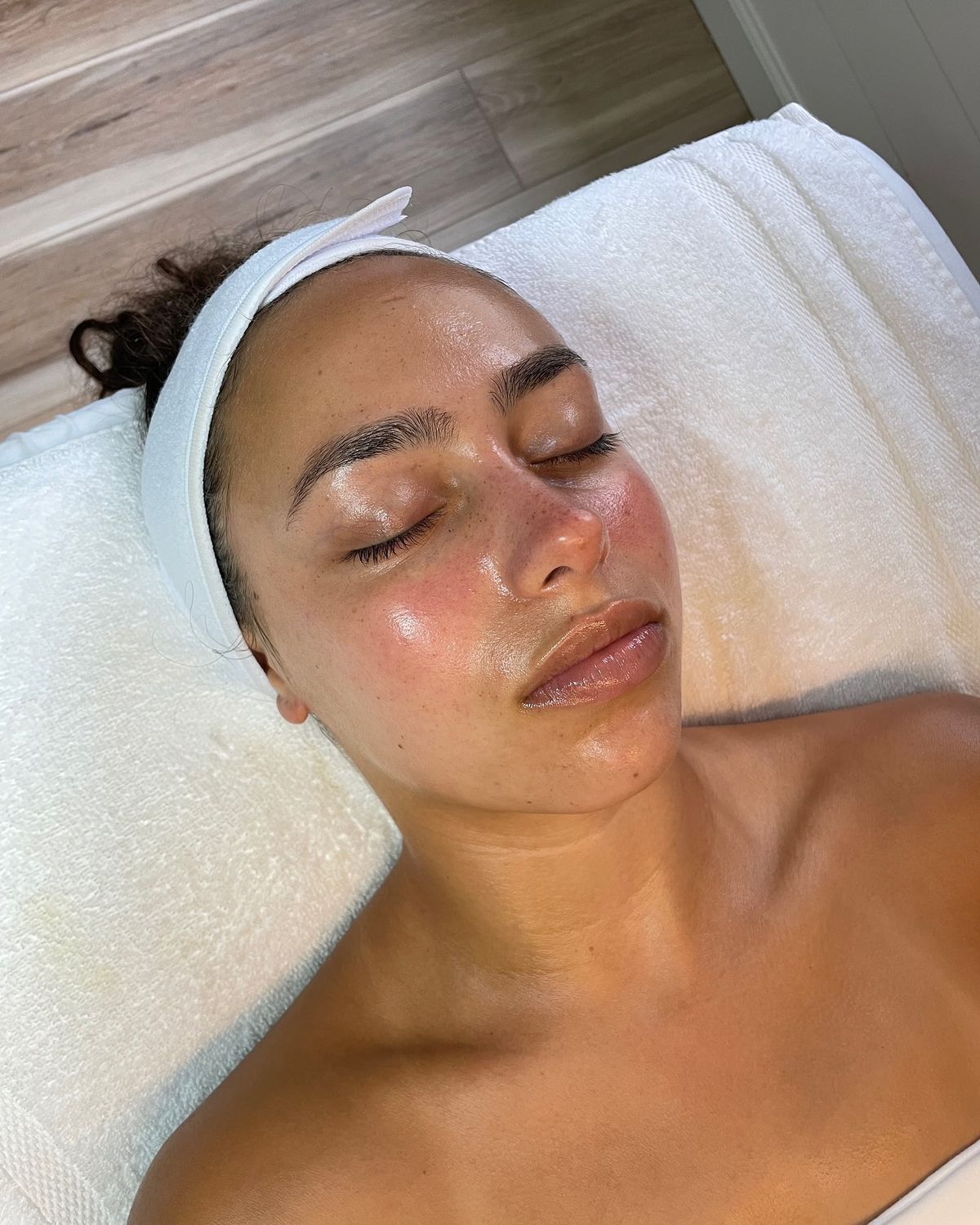 A person with eyes closed and a wet, glowing face, wearing a white spa headband while lying on a white towel.
