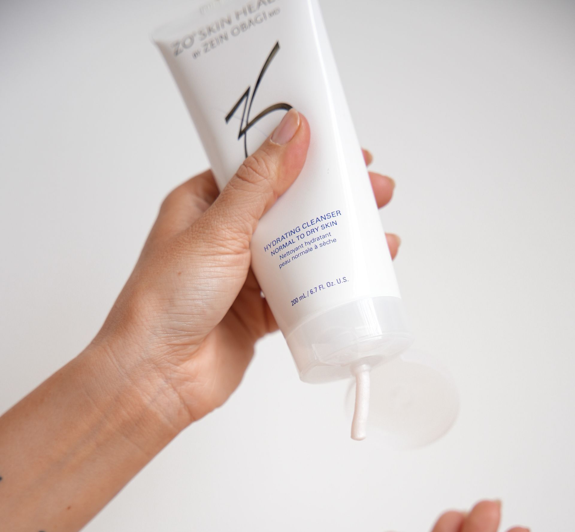 A person squeezing a tube of ZO Skin Health cream. The white cream is dispensed.