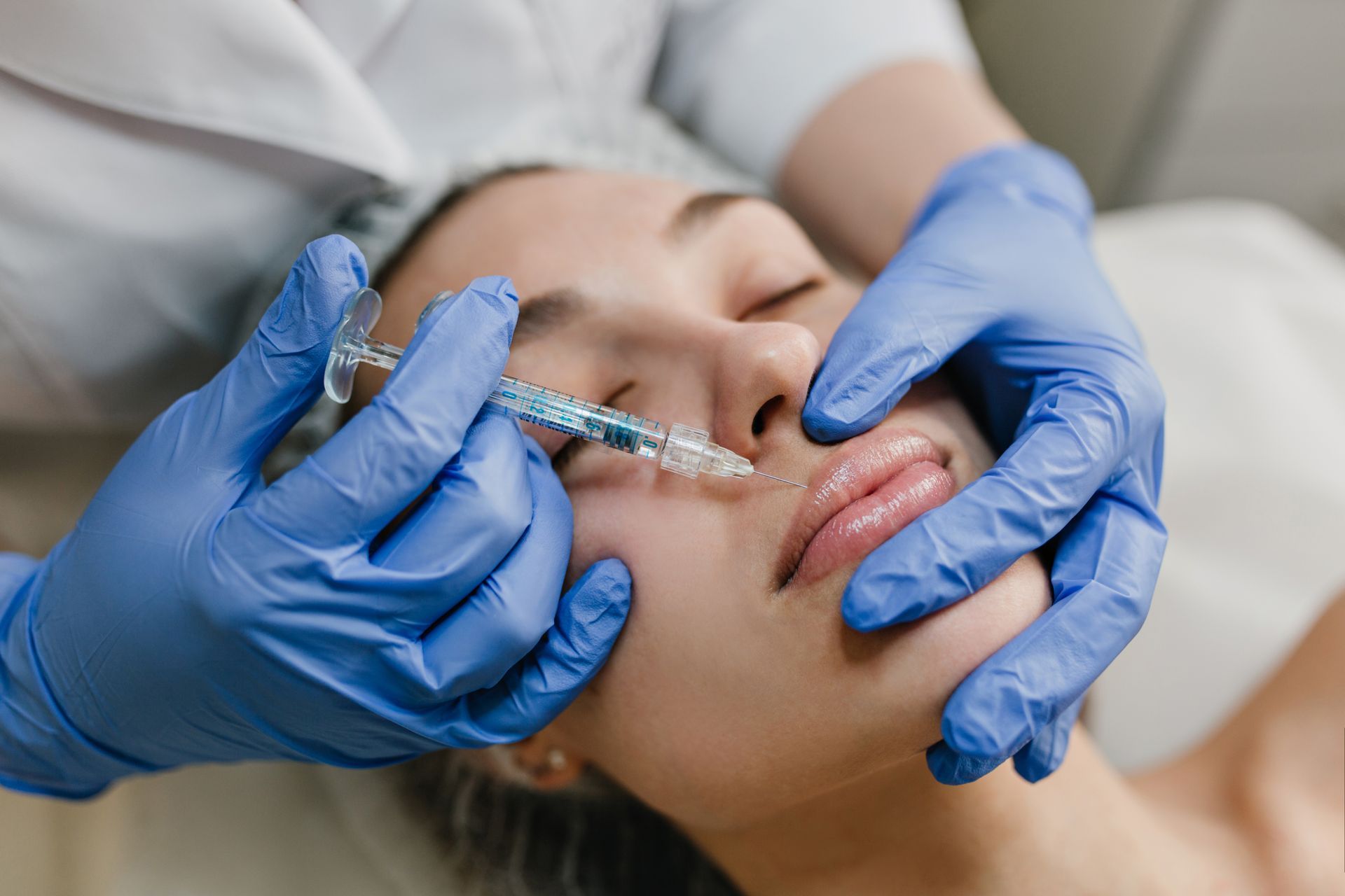 A practitioner in blue gloves uses a syringe to perform a dermal filler injection on a person’s upper lip.