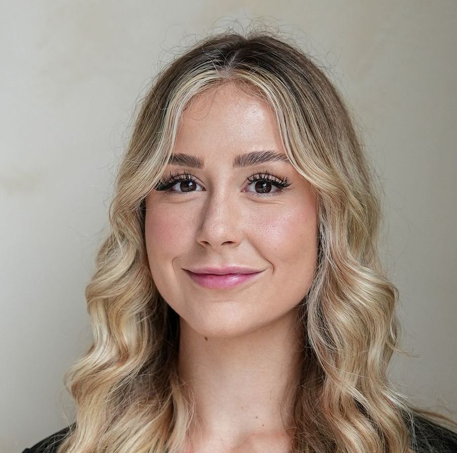 Blonde woman with wavy hair smiles. Indoors, neutral background.