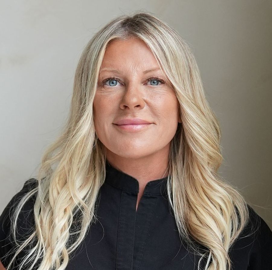 Blonde woman with blue eyes, smiling slightly, wearing a black shirt in front of a light-colored background.