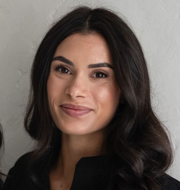 Woman with long dark hair, smiling, holding a camera on a tripod, indoors against a white background.