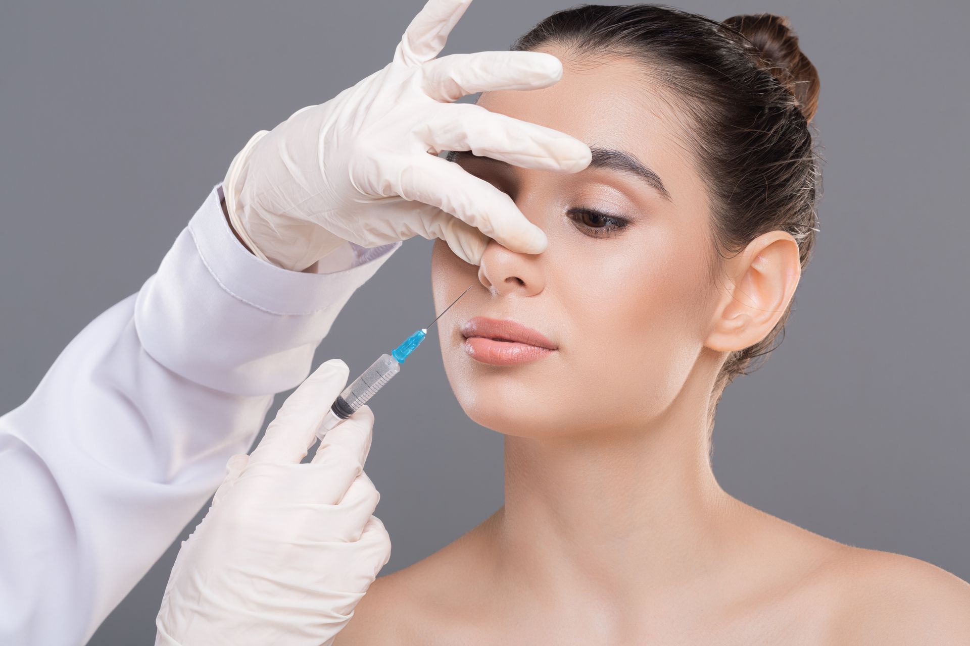 A medical professional in white gloves administers a facial injection to a person's nose against a gray background.