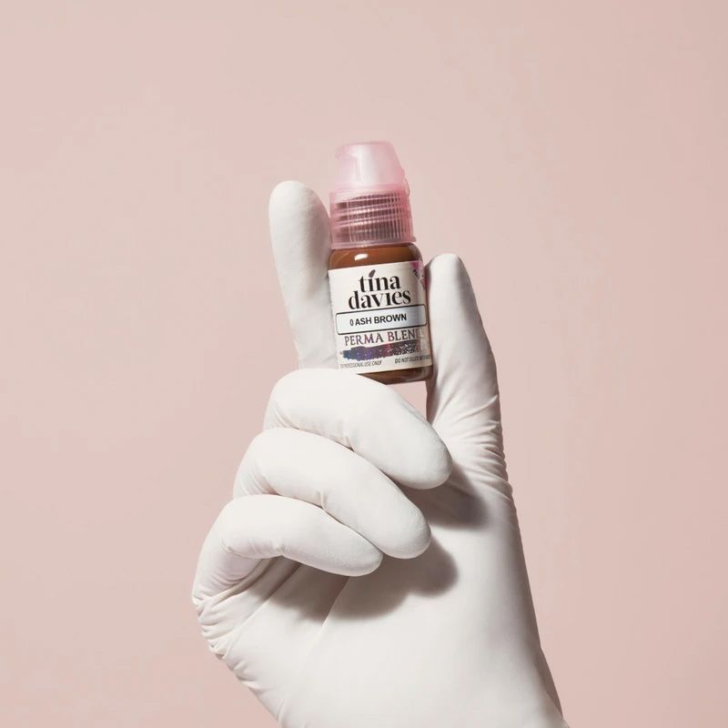 Gloved hand holding a brown, pink-capped Tina Davies brand tattoo ink bottle against a pink backdrop.