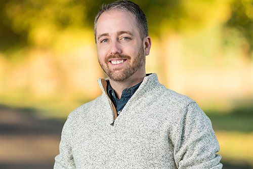 Man with light hair and beard, wearing a light sweater, smiling outdoors.