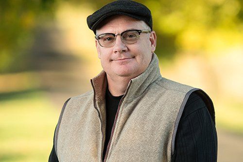 Man wearing glasses and cap, smiles outdoors, wearing vest over black sweater.