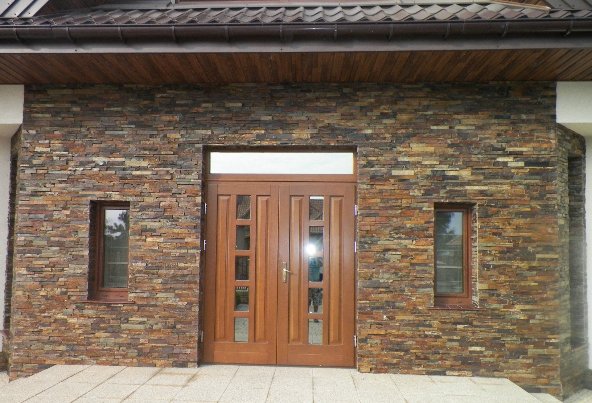 Brązowa kamienna fasada z drewnianymi podwójnymi drzwiami, oknami i okapem.