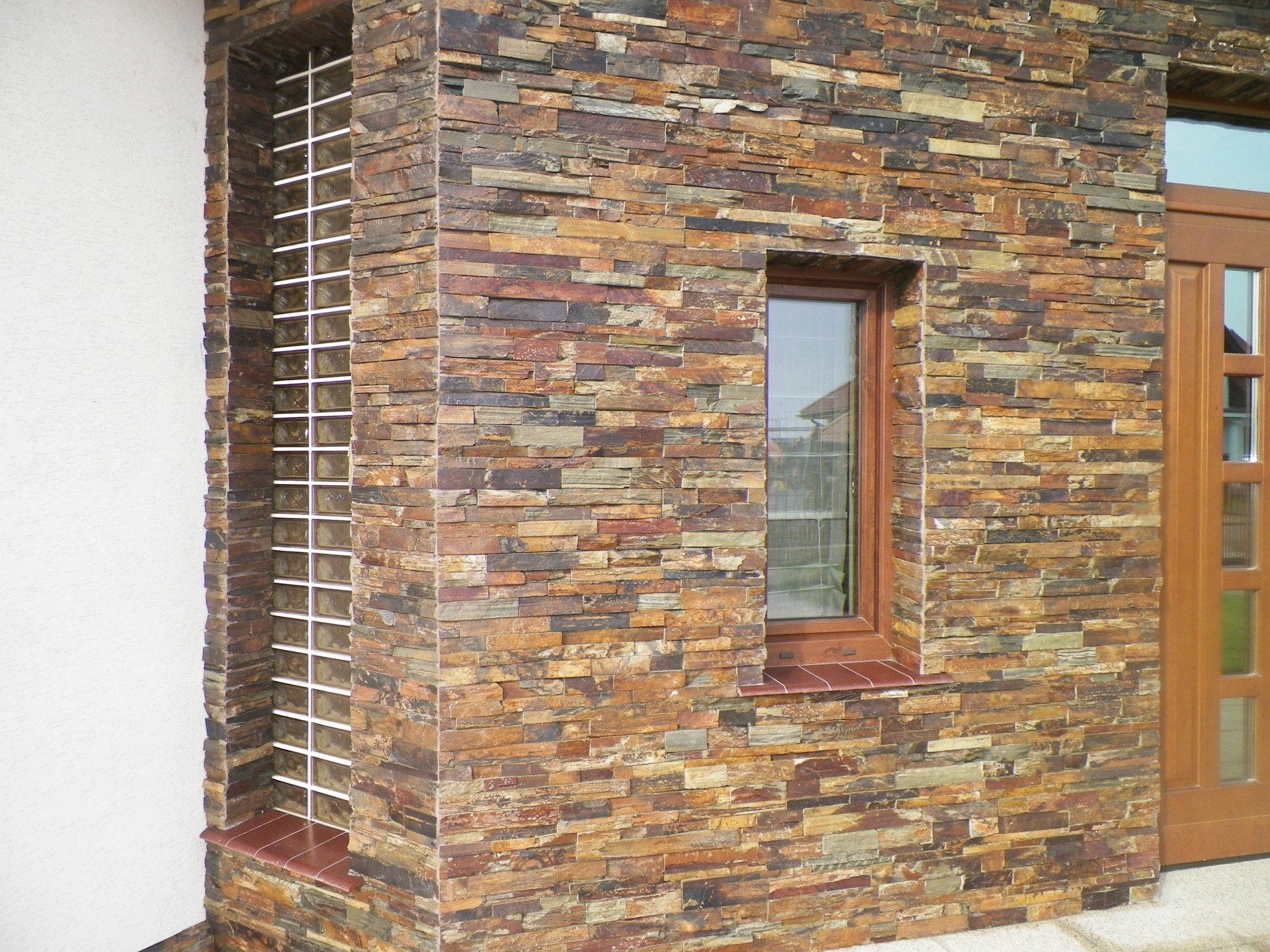 Narożnik budynku ze ścianami wyłożonymi kamieniem, małym oknem i metalową kratą.