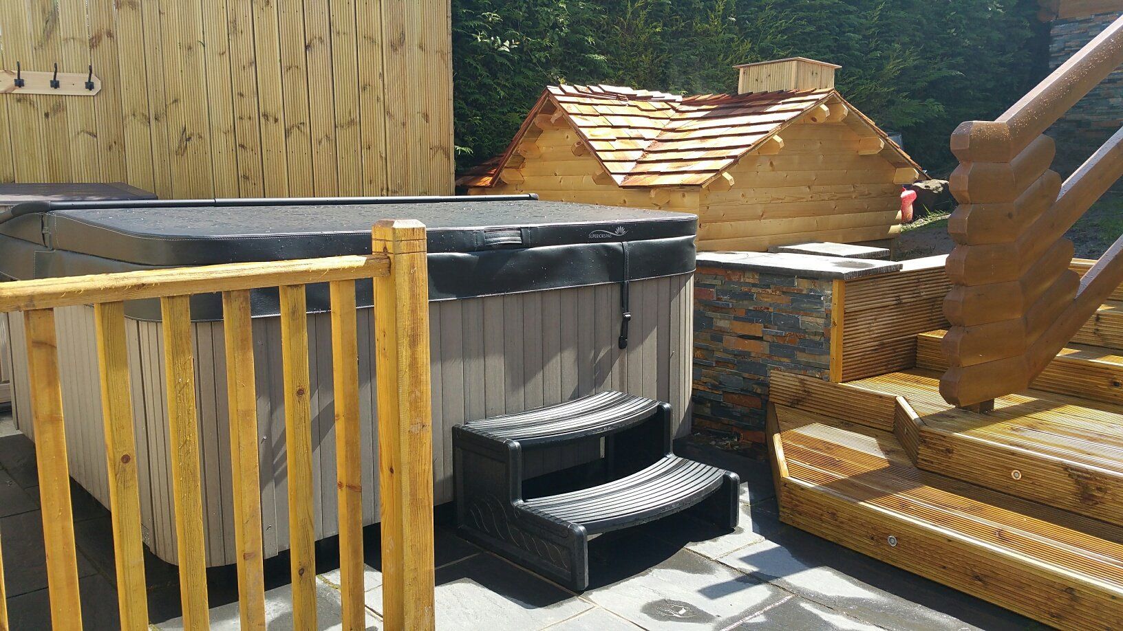 Wanna z hydromasażem z czarną pokrywą, schodami i drewnianą balustradą na tarasie. Mała drewniana konstrukcja w tle.