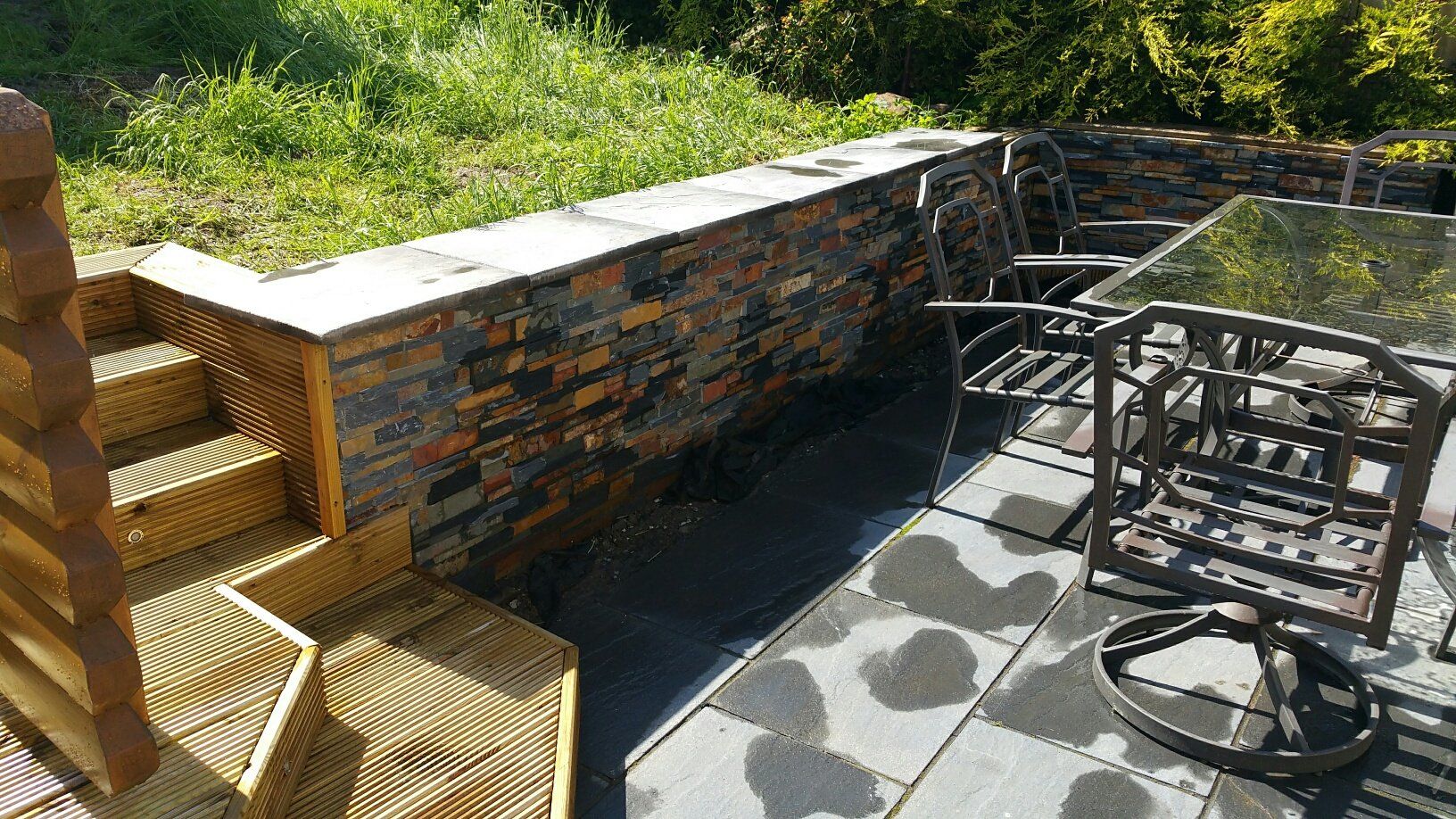 Kamienny mur i patio z meblami ogrodowymi i schodami prowadzącymi na trawiasty teren.