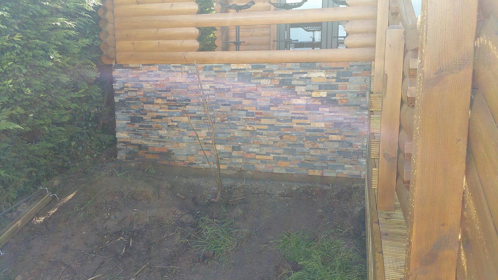 Kamienny mur pod drewnianą konstrukcją ze szczegółami z bali; zieleń i ziemia na pierwszym planie.