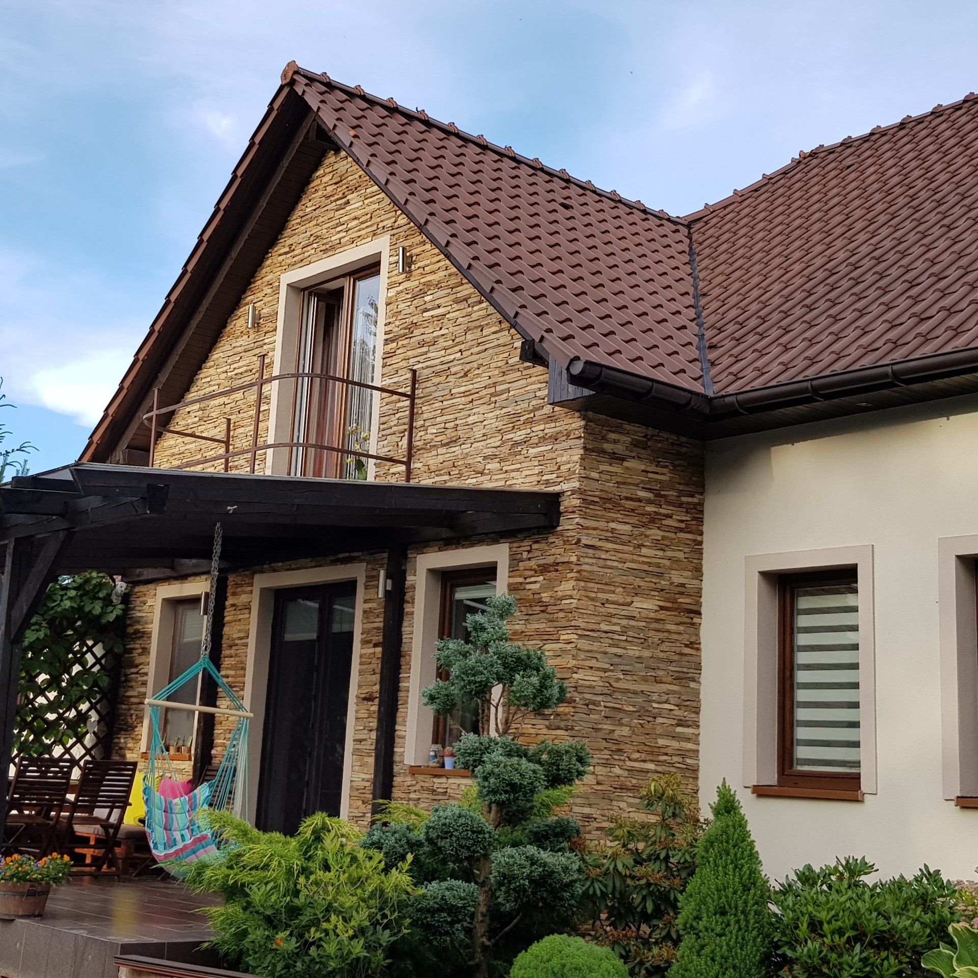 Dom z brązowym dachem i kamienną fasadą, gankiem, balkonem i ogrodem.