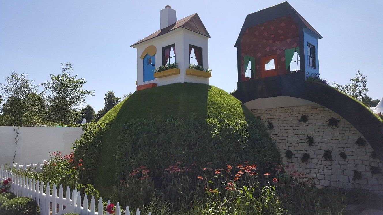 Dwa fantazyjne domy na trawiastych wzgórzach, z białym płotem na tle błękitnego nieba.