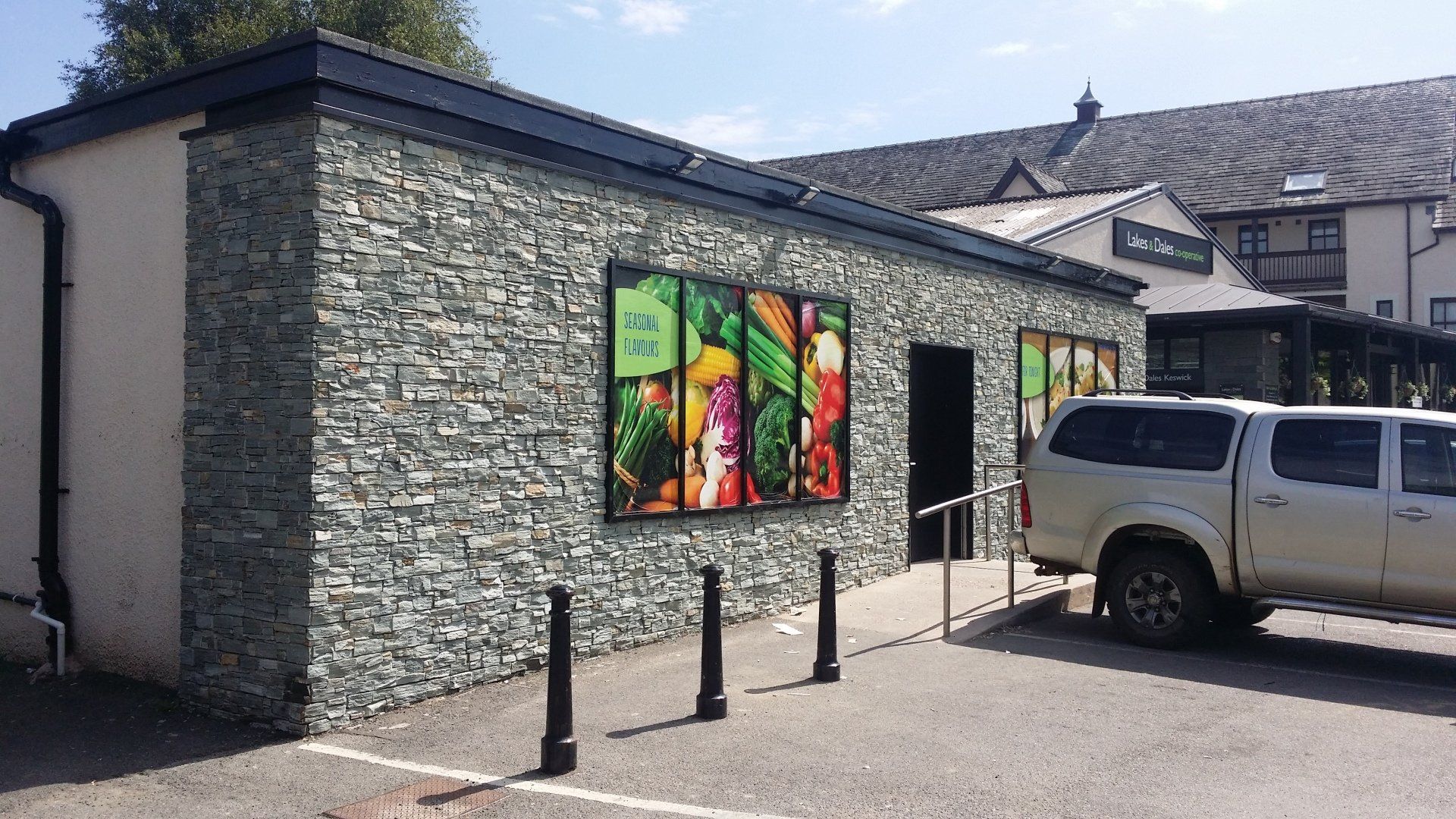 Sklep spożywczy z fasadą z teksturowanego kamienia i kolorowymi plakatami z produktami. W pobliżu zaparkowany jest srebrny samochód ciężarowy.