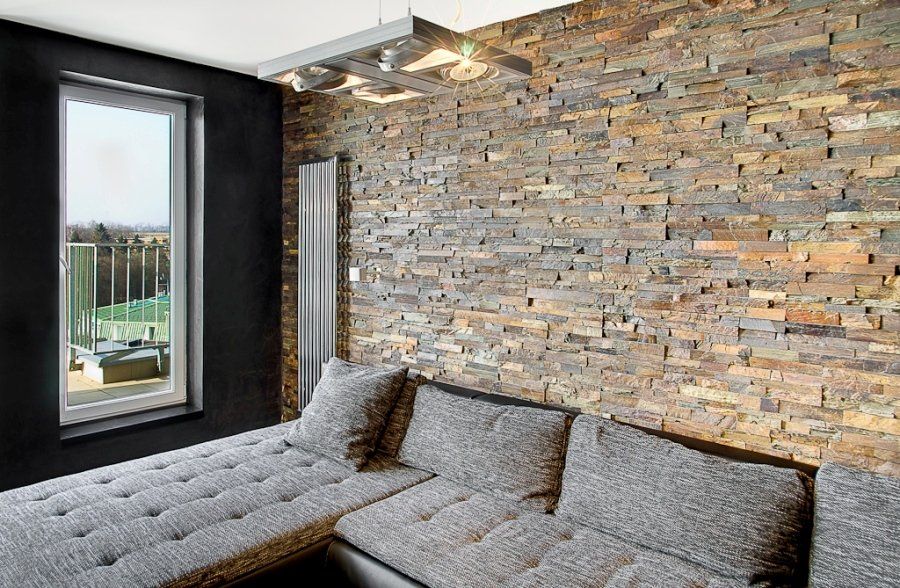 Salon ze ścianą z kamiennymi akcentami, szarą narożną sofą i oknem z widokiem na balkon.