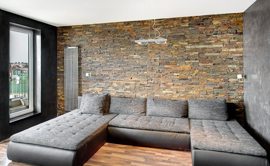 Szara kanapa w kształcie litery U na tle ściany z kamienia o fakturze drewna w nowoczesnym salonie, z ciemną ścianą po lewej stronie.