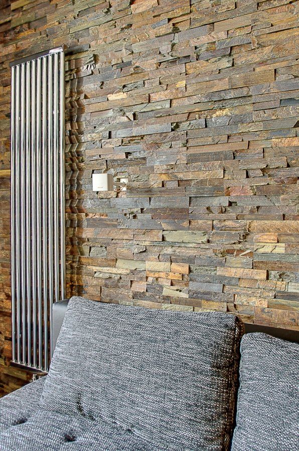 Nowoczesny srebrny grzejnik na ścianie z kamienia o fakturze kamienia obok szarej sofy.
