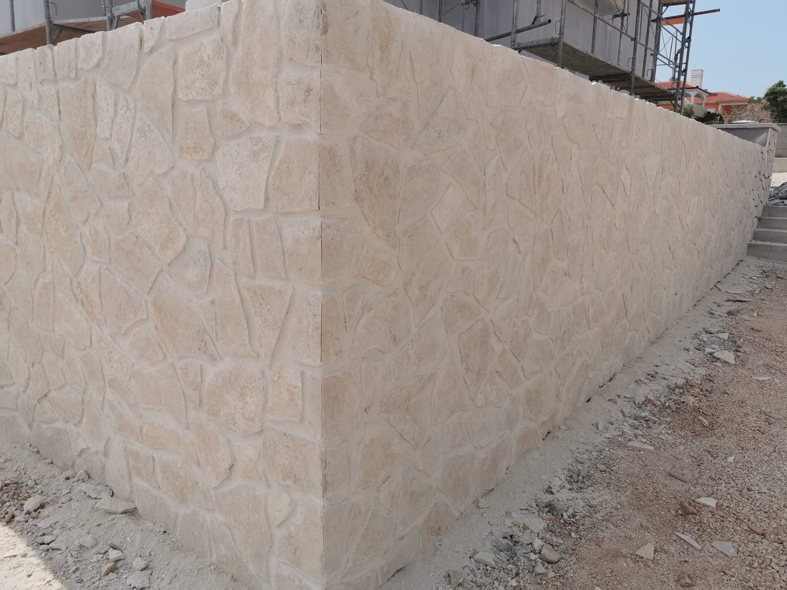 Narożnik kamiennego muru, jasnobeżowy kolor, na zewnątrz, prawdopodobnie w budowie, w pobliżu ścieżki gruntowej.